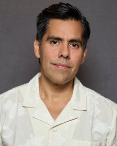 Man with dark hair, wearing a black shirt against a black background